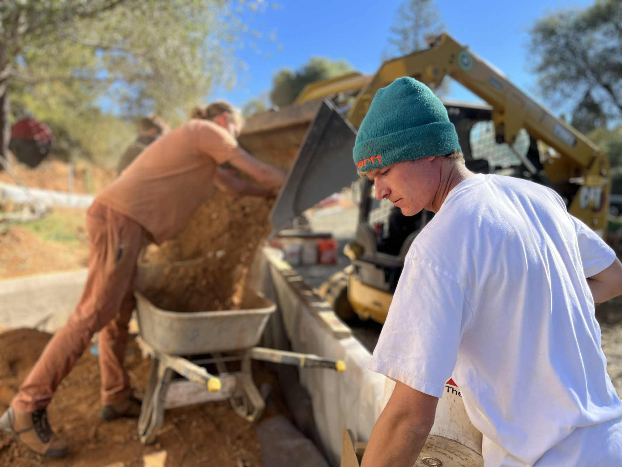 Two builders shoveling red dirt from a wheelbarrow into a trench beside a ferro-cement wall, with a skid steer behind them
