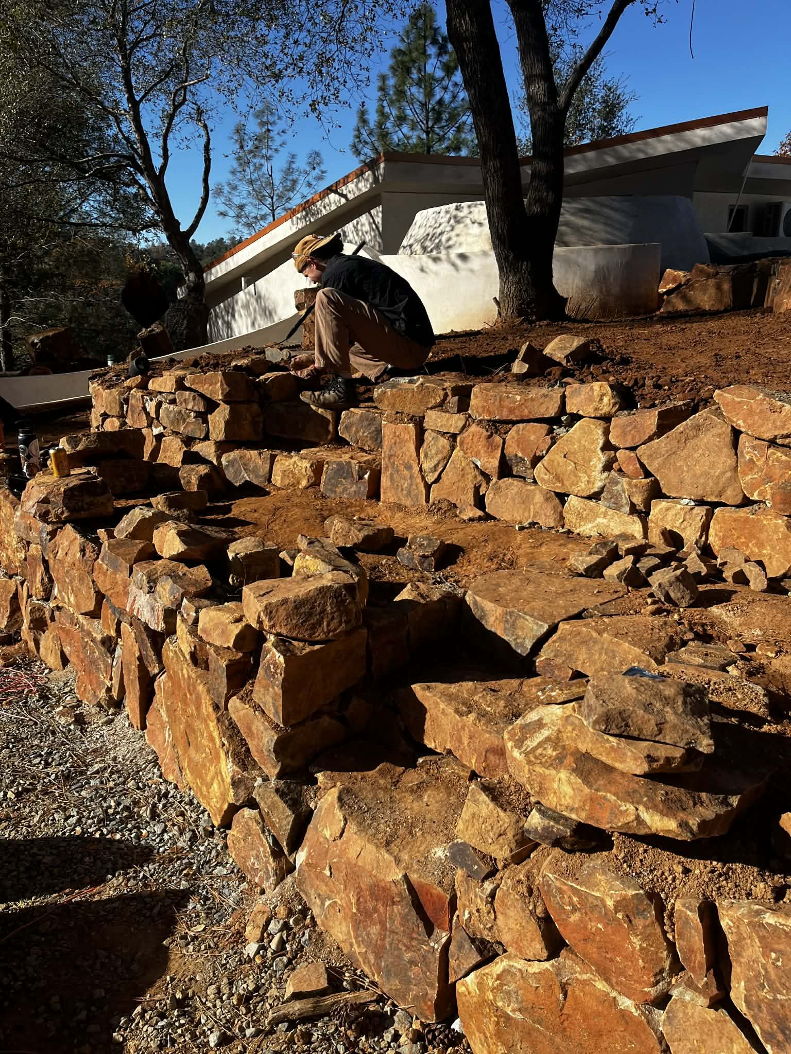 A volunteer setting rust-colored stones on a tiered dry-stacked retaining wall in front of an earth-sheltered home