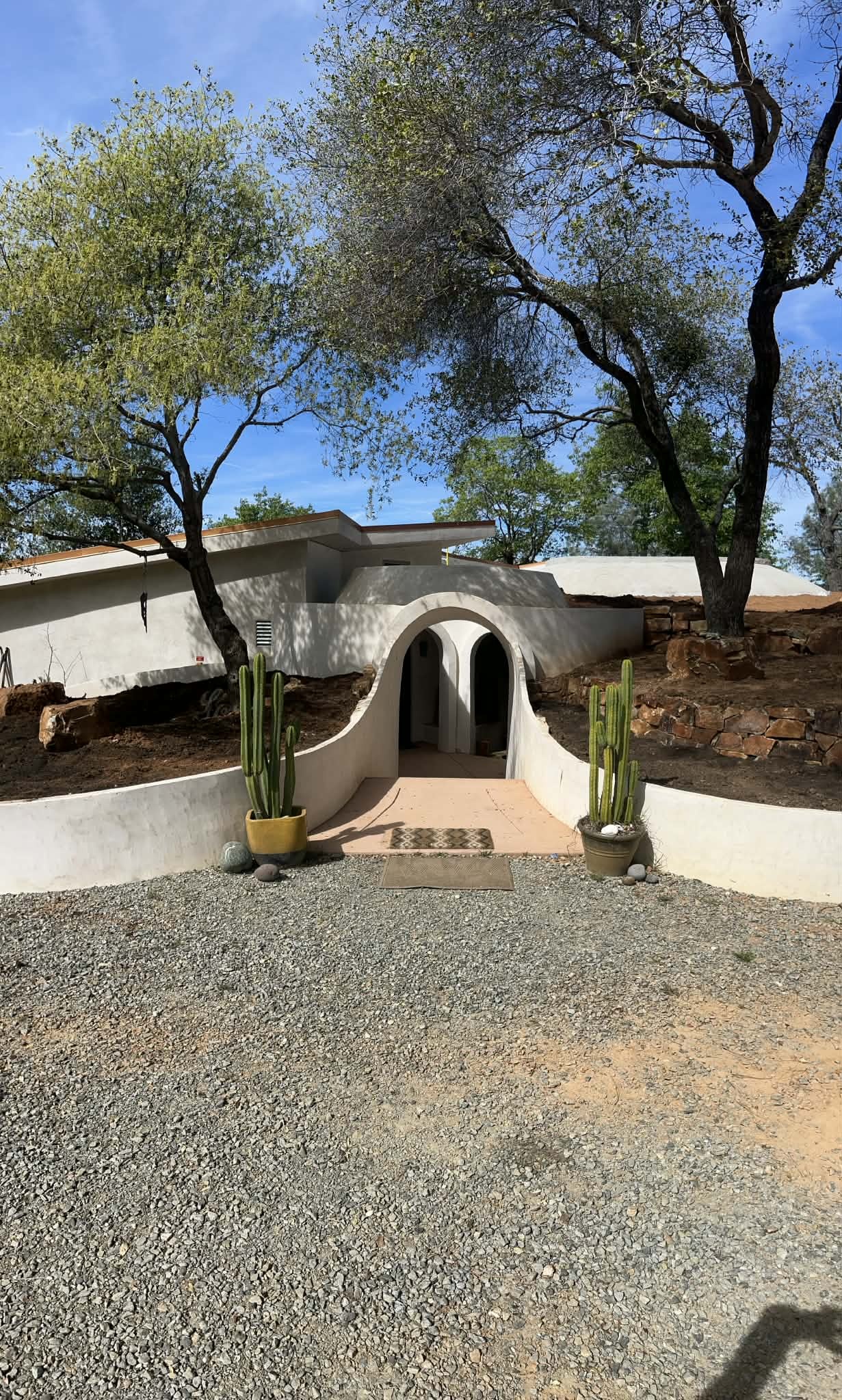 The arched white entry tunnel of an earth-sheltered home, flanked by potted cacti beneath oak trees