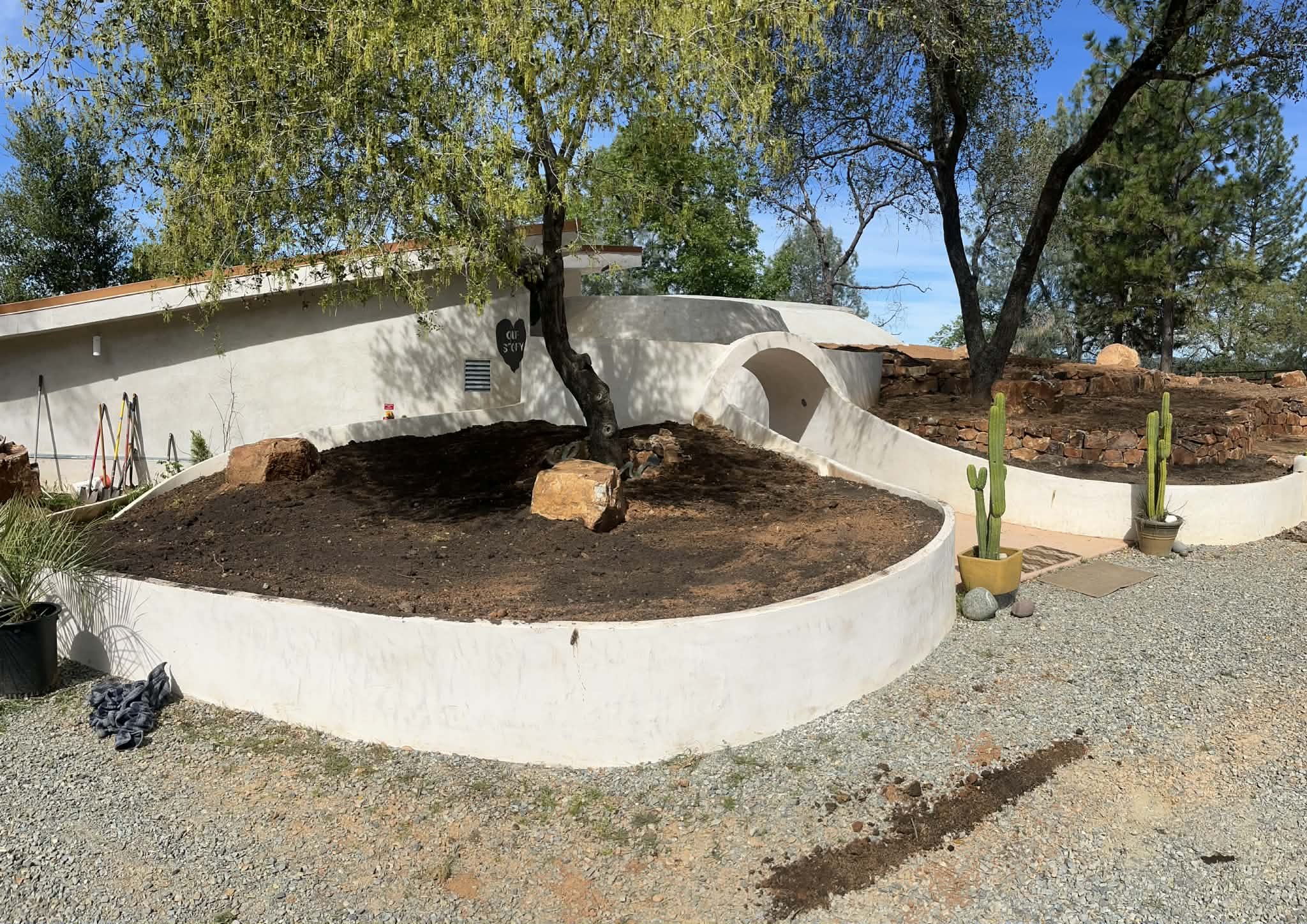 Curving white ferro-cement planter walls around an oak tree, with an arched entry and stacked stone terraces beside an earth-sheltered home