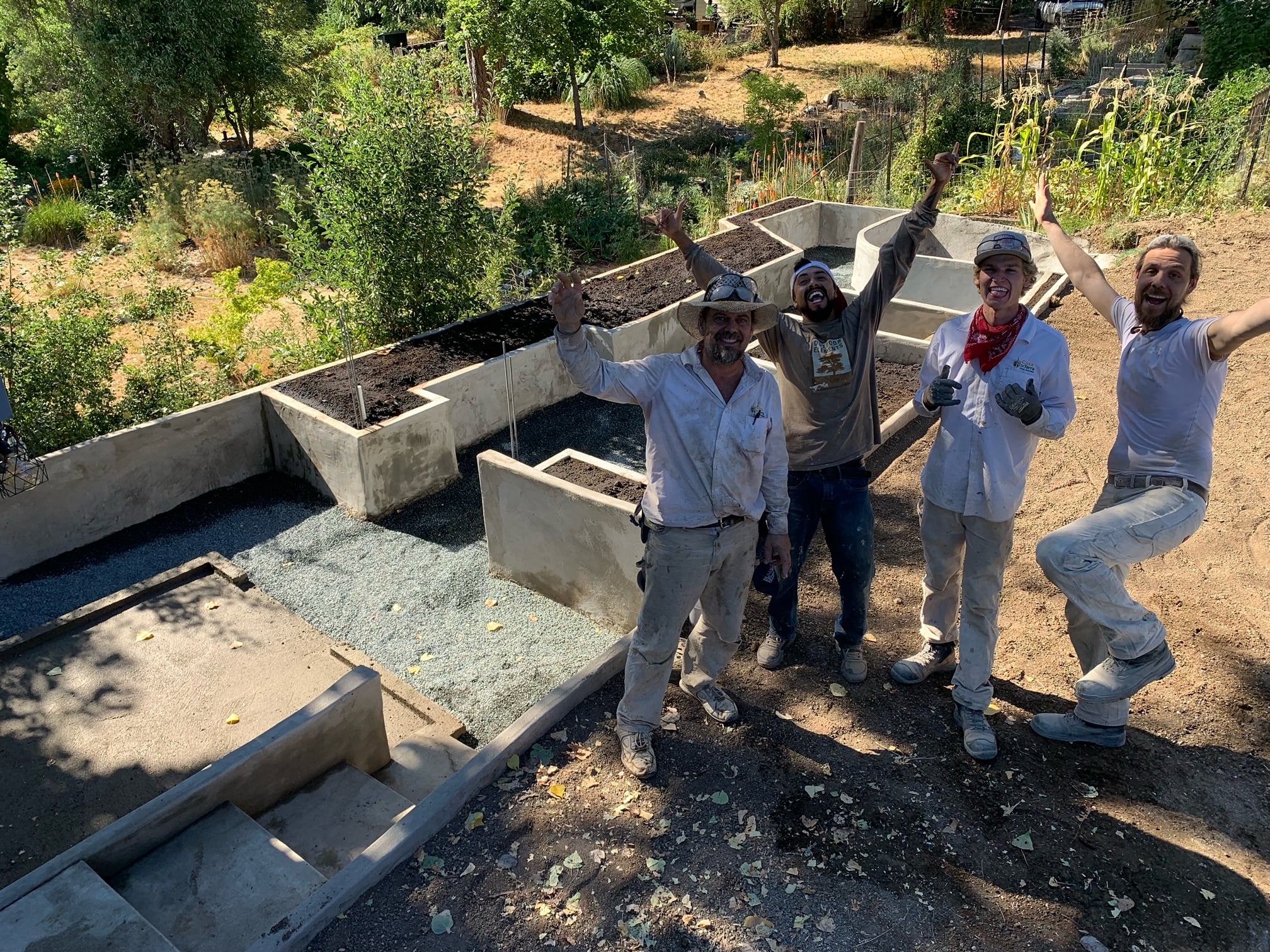 Four cheering crew members standing beside newly finished terraced ferro-cement raised garden beds filled with soil and gravel paths