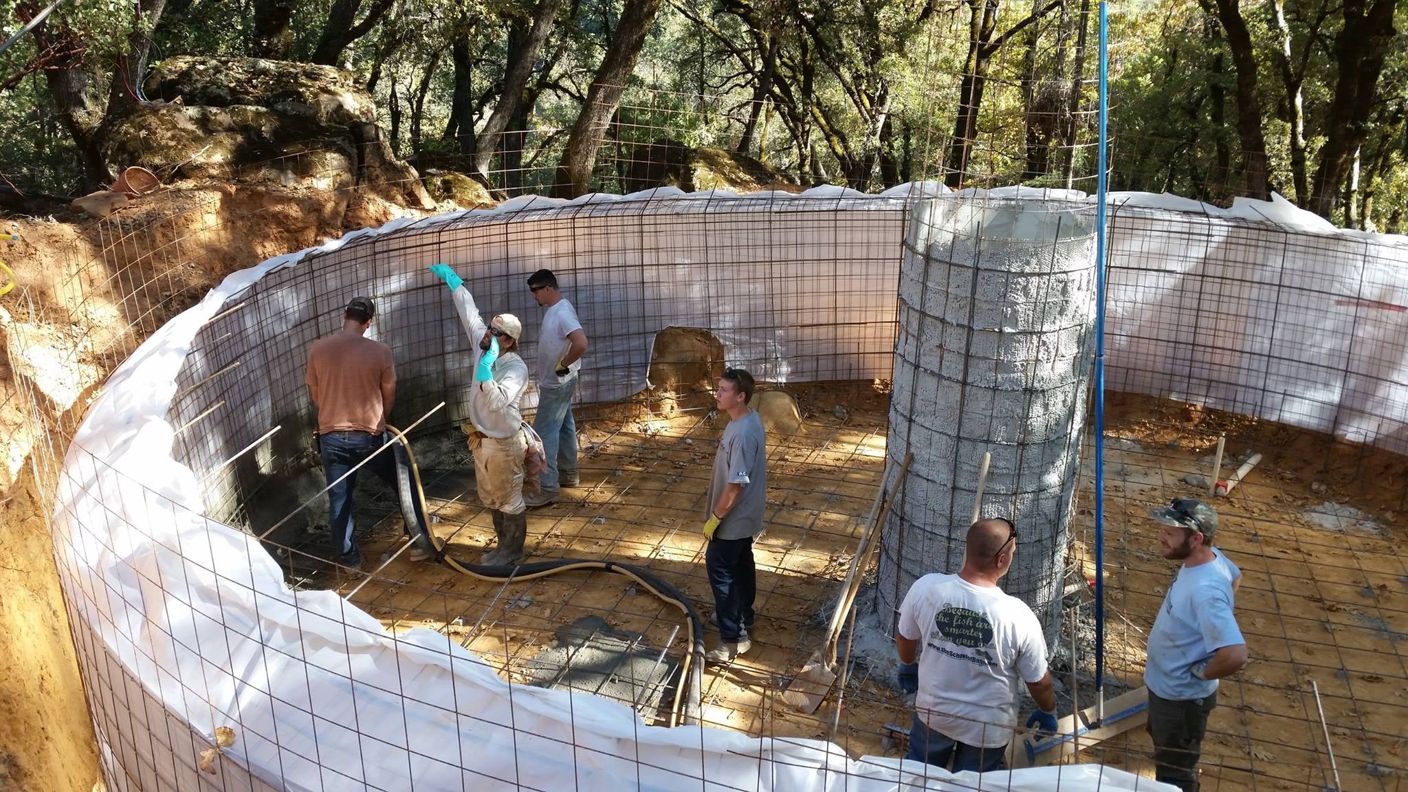 A crew spraying cement onto a large circular rebar-reinforced tank wall lined with white membrane, with a central column, in an oak forest