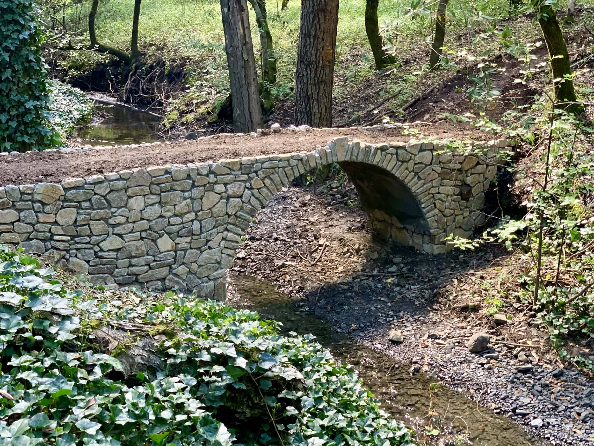 A mortared fieldstone arch footbridge spanning a shaded creek, framed by ivy and oak trunks