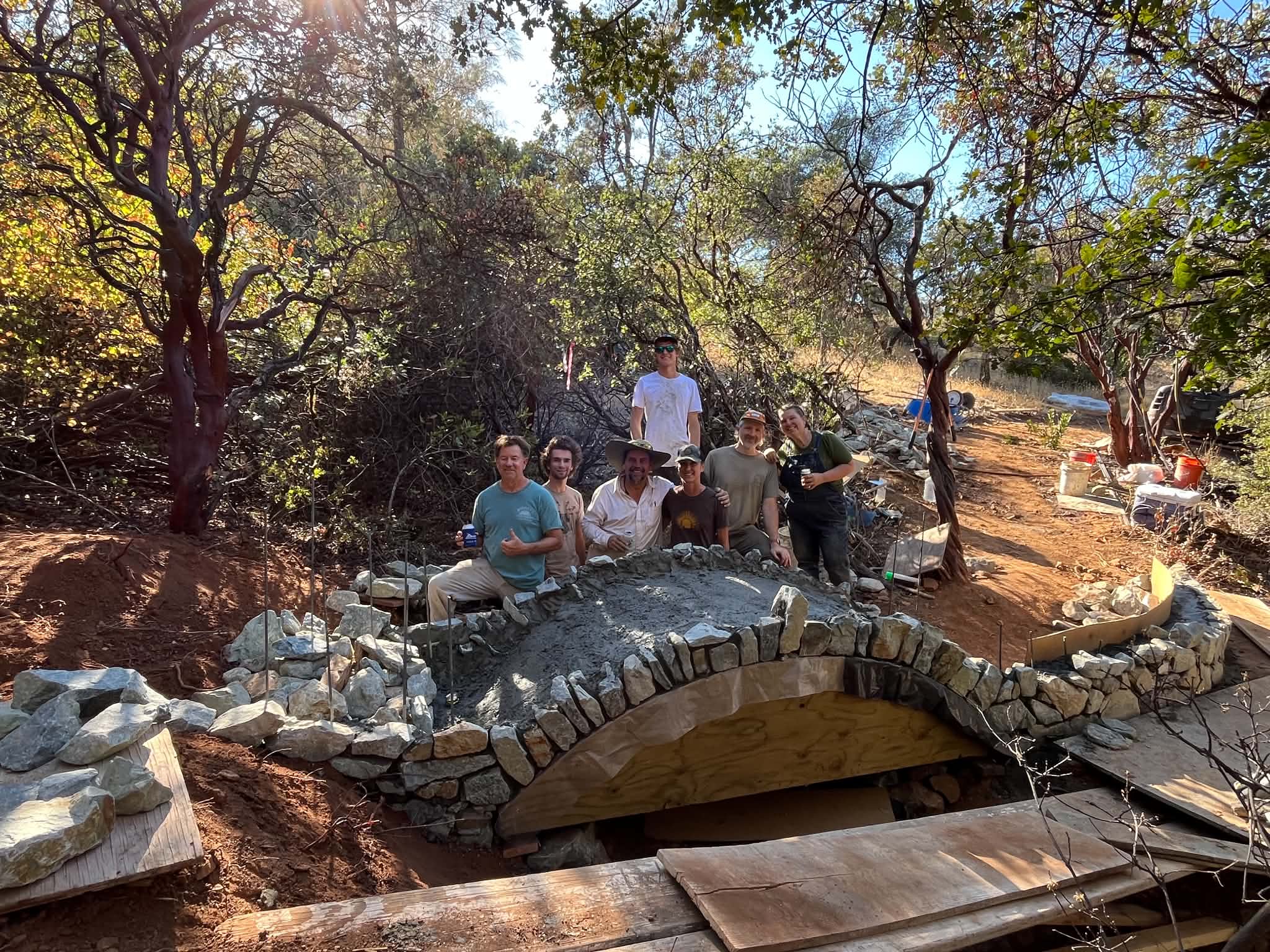 Seven Sierra Builders Guild volunteers standing behind the stone arch bridge they are building among manzanita trees, with the plywood arch form still in place