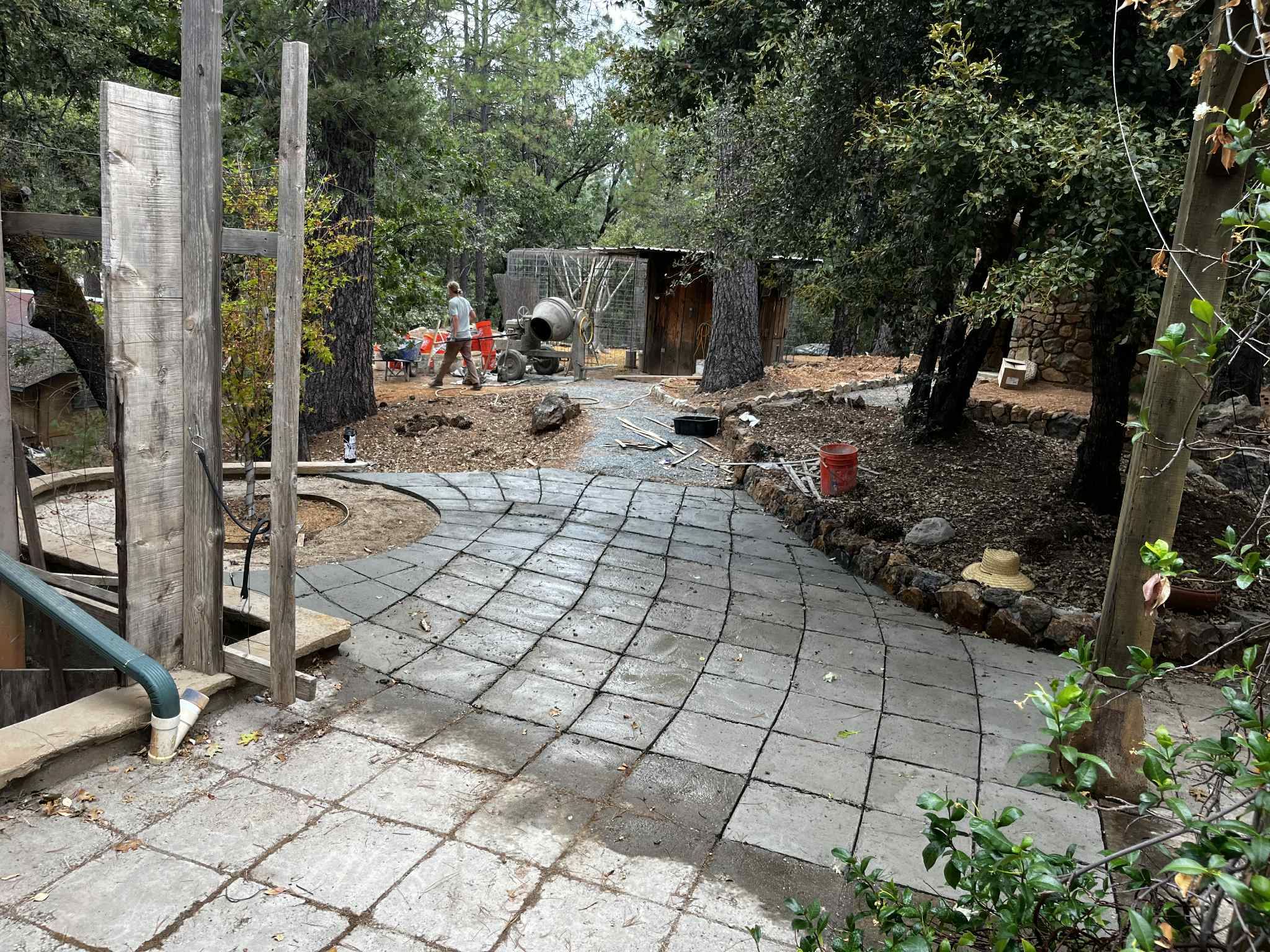 A freshly poured concrete patio scored in a curving grid, winding through a wooded yard toward a cement mixer